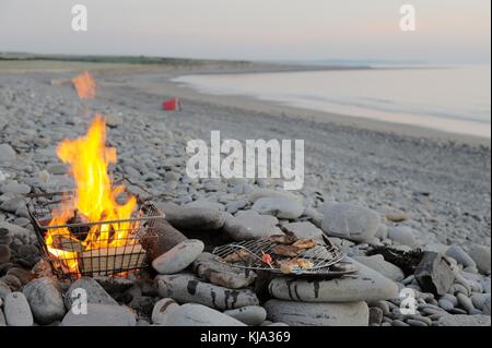 Reste einer Mahlzeit auf einem Grill in einem Wiederverwendet Warenkorb am Strand mit Zelt gekocht und Defokussierten person im Hintergrund, Wales, Großbritannien Stockfoto