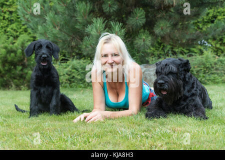 Lächelnde blonde Frau liegt zwischen Welpe und erwachsener Hund von riesigen Schwarzen Schnauzer Hund auf der Liegewiese im Garten. Stockfoto