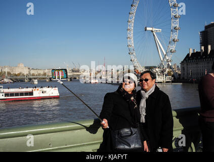 Eine asiatische Paar für einen selfie selfie mit einem Stock auf die Westminster Bridge London mit dem London Eye im Hintergrund darstellen. Stockfoto