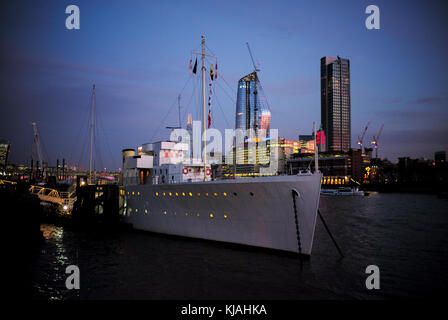 HQS Wellington günstig auf der Themse neben Victoria Embankment London in der Dämmerung mit dem Londoner Skyline im Hintergrund. Stockfoto