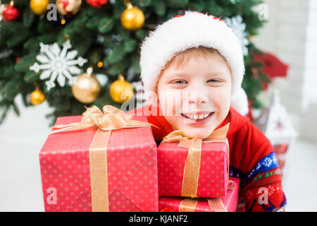 Closeup Portrait von Angeregten niedlichen Kind im Urlaub Weihnachten Innenraum. Happy adorable Kid mit viele Geschenke in rot Geschenkpapier posieren. horiz. Stockfoto