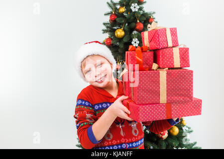 Closeup Portrait von Glücklich lächelnde Kind holding Haufen von vorhandenen Boxen in den Händen bereit, um sie zu öffnen. junger Mann, Santa Hat und Red Pyjama. Urlaub gree Stockfoto