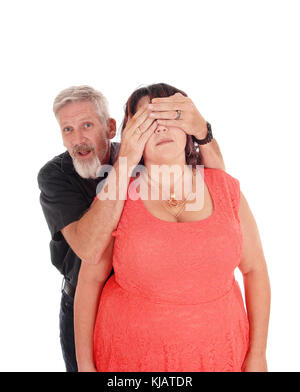 Ein Paar mittleren Alters steht für weissen Hintergrund und der Mann hält seine Hände über die Augen seiner Frau Stockfoto
