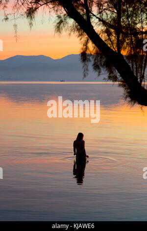 Eine einsame Frau Badegast wird reflektiert, wie sie den ruhigen Wasser der Ägäis. Sie ist gegen die flammenden Farben einer Dämmerung Himmel. Stockfoto