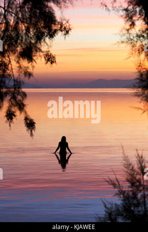 Eine einsame Frau Badegast wird reflektiert, wie sie den ruhigen Wasser der Ägäis. Sie ist gegen die flammenden Farben einer Dämmerung Himmel. Stockfoto