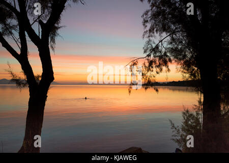 Das ruhige Meer spiegelt das helle orange Dämmerung Himmel und plätschert der fernen, silhouetted Kopf eines einsamen Schwimmer umgeben. Stockfoto