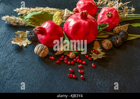 Rote reife Früchte Granatapfel mit Nüssen und Beeren, trockene Blätter und Zweige, auf einem schwarzen Holz- Hintergrund Stockfoto