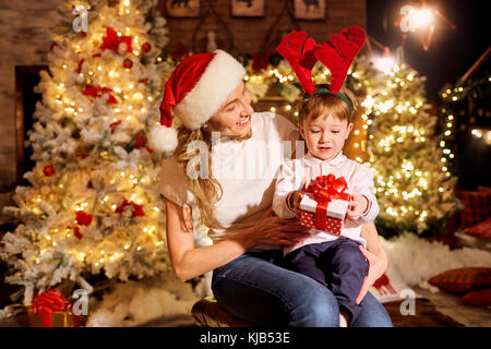 Mama gibt ein Geschenk in einer Box zu ihrem Sohn in einem Zimmer am Weihnachtstag. Stockfoto