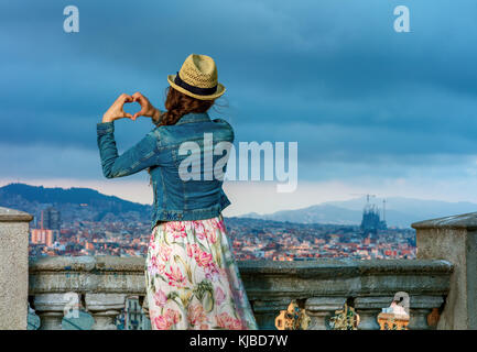 Perfekter Abend mit atemberaubenden Blick. Hinter der eleganten reisenden Frau im langen Kleid und Strohhut in Barcelona, Spanien, herzförmige Hand Stockfoto