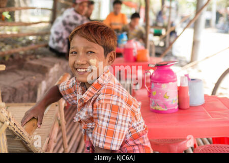 Ein junger Burmesjunge mit einem Lächeln und Ingwerhaaren und traditionellen Thanaka-Dekorationen im Gesicht sitzt in einem lokalen Straßencafé auf dem Pyay-Markt Stockfoto