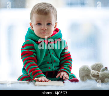 Kleinen Jungen in roten und grünen Strampler mit Teddy sitzt auf den Hintergrund des Fensters. Stockfoto