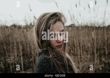 Junge und schöne Frau mit langen blonden Haaren in Wolle Mantel bekleidet den Blick über ihre Schulter. Mädchen stehen auf rohrkolben Hintergrund. Outdoor. Stockfoto