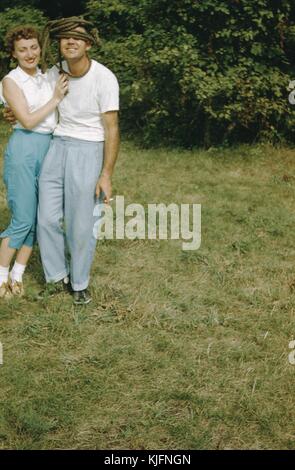 Ein Foto von einem jungen Mann und einer Frau, die zusammen posieren, sie sind in einer verspielten Umarmung, der Mann hat ein dickes Seil um den Kopf und die Augen gewickelt, er raucht auch eine Zigarette auf dem Foto, sie stehen in einem grasbewachsenen Gebiet mit Bäumen dahinter, 1952. Stockfoto