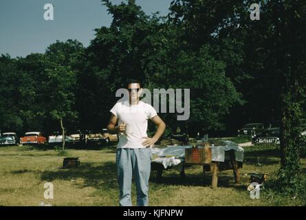 Ein Foto von einem Mann mit einem Becher posiert, der Mann trägt ein weisses T-Shirt und blaue Hosen, hinter ihm ein Picknick Tabelle zu sehen in einem Tuch bedeckt und angehäufte Höhe mit Elementen für ein Picknick, die Umgebung ist ein Park mit Gras und Bäume, viele Oldtimer gesehen kann im Hintergrund des Fotos, 1952 abgestellt werden. Stockfoto