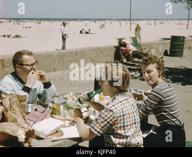 Drei Personen, ein Mann und zwei Frauen, sitzen an einem Tisch, auf einem Betonweg neben dem Strand, essen eine große Menge Essen und trinken ein alkoholfreies Getränk, im Hintergrund liegen Menschen auf dem Sand am Strand, 1952. Stockfoto