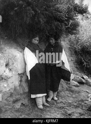 Ein Foto von zwei Hopi-Frauen, die lange schwarze Kleider tragen und Decken über den Schultern hängen, sie stehen auf einem Fels- und Schlammweg, der unter einem Baum in einen Hügel geschnitten ist, 1906. Aus der New York Public Library. Stockfoto