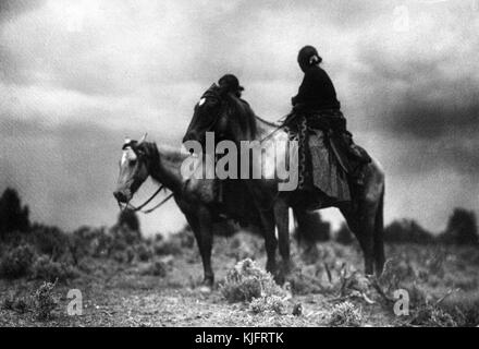 Eine Fotografie von zwei Navajo-Frauen zu Pferd, die Frau im Hintergrund wird vom Pferd im Vordergrund verdeckt, die Frau im Vordergrund trägt ein dunkles Oberteil und einen langen gemusterten Rock, sie stehen unter kurzem Wüstengras mit stürmischem Himmel als Hintergrund, 1906. Aus der New York Public Library. Stockfoto