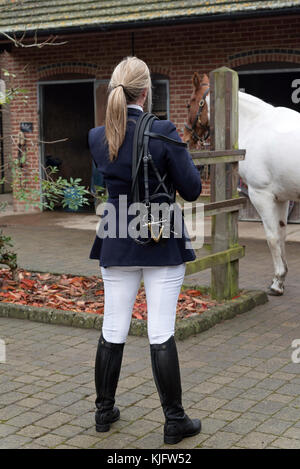 Blonde Reiterin, die im Stallgarten mit einem Zaumzeug über ihrer Schulter steht. November 2017 Stockfoto