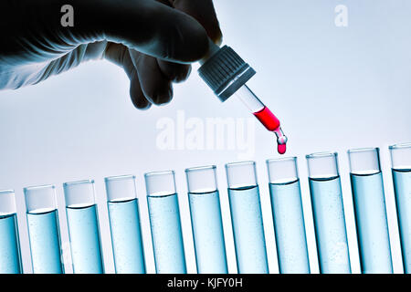 Hand eines Forschers mit Latex Handschuh fallen rote Lösung mit tropfer in einer Reihe von Reagenzgläsern gefüllt mit blauer Flüssigkeit. Ansicht von vorn Stockfoto