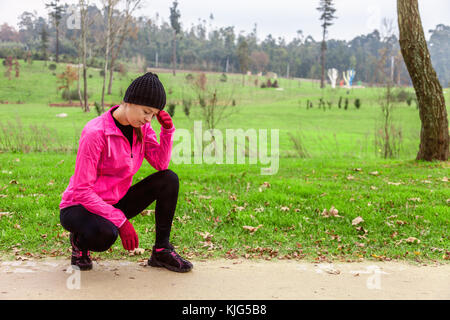Junge Frau Schwindelgefühl oder mit Kopfschmerzen nach dem Zug an einem kalten Wintertag auf der Spur eines städtischen Park. weiblichen Athleten mit Rosa Stockfoto