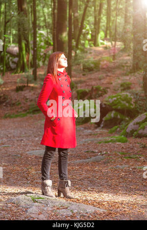 Frau genießen die Wärme im Winter Sonnenlicht allein auf einem forest park Weg einen roten Mantel Stockfoto