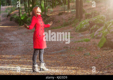 Frau genießen die Wärme im Winter Sonnenlicht allein auf Forest Park weg mit offenen Armen empfangen von Lichtstrahlen, trug einen roten Mantel oder Jacke Stockfoto