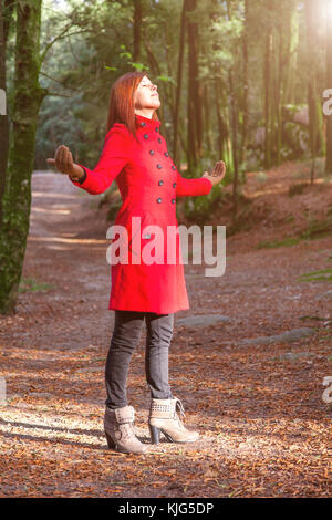 Frau genießen die Wärme im Winter Sonnenlicht allein auf Forest Park weg mit offenen Armen empfangen von Lichtstrahlen, trug einen roten Mantel oder Jacke Stockfoto