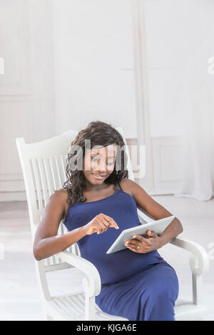 Schöne afrikanische schwangere Frau ist mit einem digitalen Tablet beim Sitzen auf dem Stuhl Stockfoto