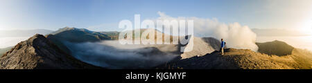 Panorama des inneren Vulkankraters Gunung Bromo mit Schwefeldampf und Morgennebel bei Sonnenaufgang in Java, Indonesien, mit einem männlichen Erwachsenen. Stockfoto