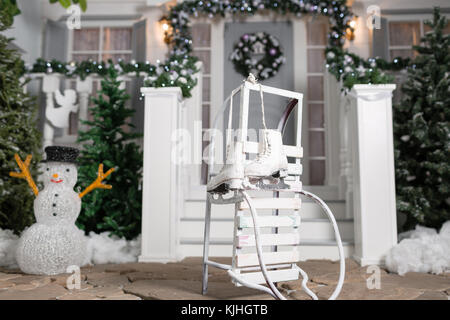 Schlitten und Schlittschuhe im Vordergrund. Haus Eingang für Feiertage eingerichtet. Weihnachten Dekoration. girlande der Tannenbaum, Zweige und Lichter auf dem Geländer Stockfoto