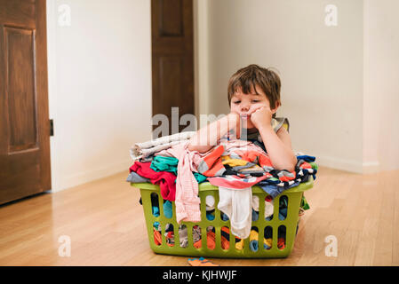 Müde Junge sitzt auf dem Boden lehnte sich auf der Wäsche Korb gefüllt mit Kleidung Stockfoto