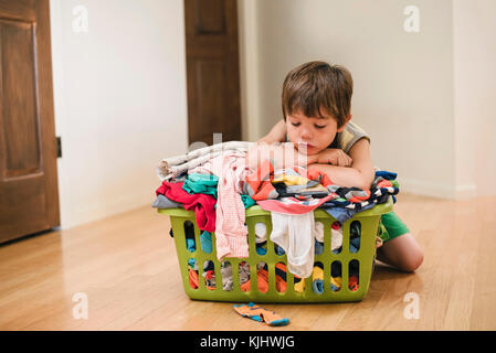 Müde Junge sitzt auf dem Boden lehnte sich auf der Wäsche Korb gefüllt mit Kleidung Stockfoto