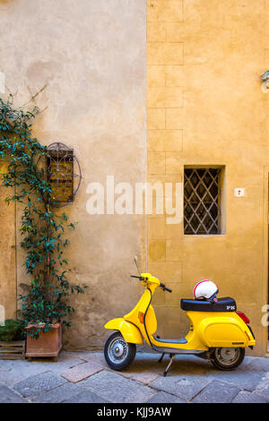 Im Vintage-Stil, Gelbe Vespa mit Helm auf dem Sitz, geparkt vor einer zweifarbigen gelben Steinmauer, die im Dorf Pienza in der Toskana aufgenommen wurde. Keine Personen Stockfoto