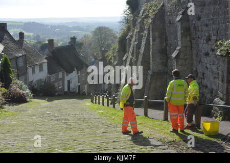 Drei Arbeiter diskutieren, wie sie das Gras von den Kopfsteinpflaster von Shaftesbury Gold Hill, Dorset, England, räumen Stockfoto