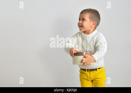 Eine aufgeregte lächelnde Junge in einer weißen Pullover und gelbe Jeans ist ein Geschenk von einem Karton mit einer Schleife gebunden. auf weißem Hintergrund isolieren. Christus Stockfoto