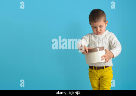 Ein wenig aufgeregt Junge in einer weißen Pullover und gelbe Jeans hält ein Geschenk. Kind auf der Suche von einem Karton mit einer Schleife gebunden. Auf Azure isolieren b Stockfoto