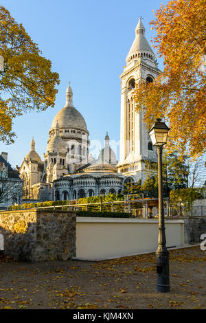 Ansicht der Rückseite der Basilika der Heiligen Herzen von Paris bei Sonnenaufgang im Herbst mit einem Vintage Style Lamp Post und orange Blätter gegen den blauen Himmel. Stockfoto