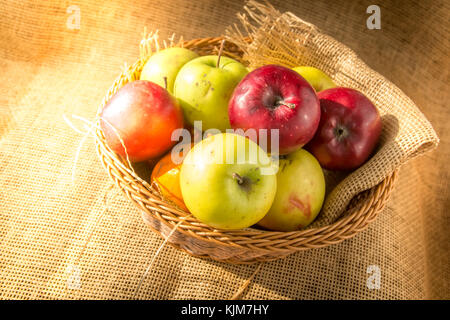 Grüne und rote Äpfel in einem Korb auf sackleinen Hintergrund mit Morgensonne leuchten. Stockfoto