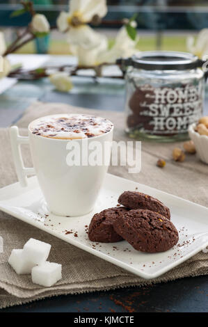 Rustikale cookies mit Kakao und Pistazien auf weißen Fach, Keramik foam Cup mit Cappuccino und Kakao Pulver, Würfelzucker, Tuch Tischdecke und andere Kit Stockfoto