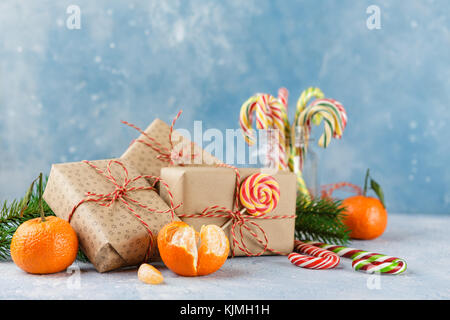 Geschenkboxen in Kraftpapier, frische Tangerinen mit Blättern, Zuckerstangen und ein Zweig der Weihnachtsbäume auf einem blauen Hintergrund Stockfoto