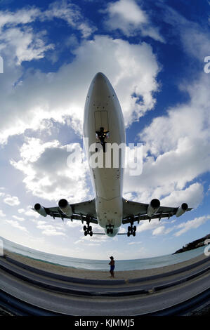 Ein Mann in einem Hut kauern wie eine A340 Pässe Overhead auf niedrigen Endrunde - Ansatz der Landung über das Meer bei Maho Beach mit Wolken Stockfoto