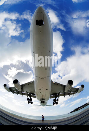 Ein Mann in einem Hut kauern wie eine A340 Pässe Overhead auf niedrigen Endrunde - Ansatz der Landung über das Meer bei Maho Beach mit Wolken Stockfoto