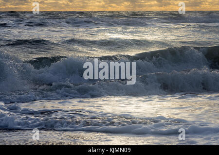 Kleine Wellen in einer unruhigen Meer rollen am Ufer mit Schaum und Gischt unter dem bewölkten Himmel und die Strahlen der Sonne Stockfoto