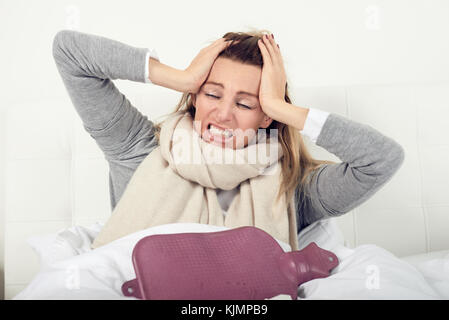 Porträt einer kranken Frau im Bett sitzend mit einer Wärmflasche beim Erfahren akute Kopfschmerzen durch Grippe oder andere schmerzhafte Krankheit verursacht Stockfoto