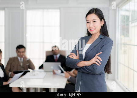 Happy Geschäftsfrau stehend mit gekreuzten Armen im Büro, während Kollegen diskutieren im Hintergrund. Stockfoto