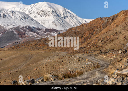Ein malerischer Herbst Landschaft mit einer felsigen Schmutz der Straße auf die Berge bedeckt mit Schnee auf dem Hintergrund der blauen Himmel Stockfoto