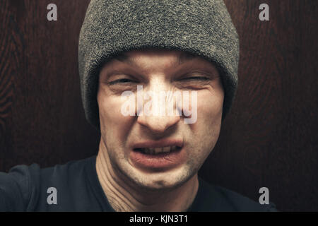 Junger Mann in grauen Hut hässliches Gesicht machen. close-up Studio Gesicht portrait auf dunklem Holz wand hintergrund, selektiver Fokus Stockfoto