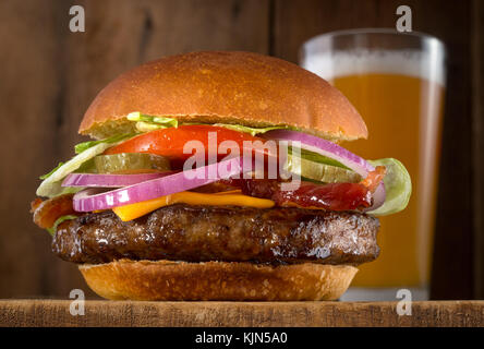 Ein saftig leckeres Cheeseburger mit Speck, Salat, Tomaten, roten Zwiebeln und Gurke mit einem Glas Bier. Stockfoto