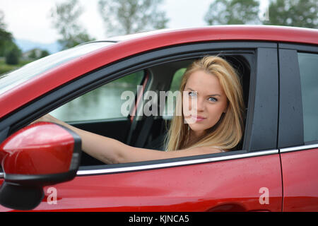 Schöne junge blonde Mädchen fahren Sie langsam und vorsichtig Brandneue rotes Auto und Suchen durch offene Fenster Stockfoto