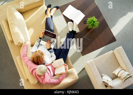 High Angle Shot eines älteren Paares zu Hause sitzen und entspannen auf dem Sofa. ältere Frau mit Laptop während der alte Mann ein Buch lesen. Stockfoto